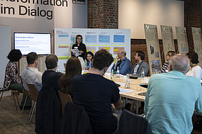 Gruppe von Teilnehmenden des vierten Workshops zum Thema Circular sitzen an einem Tisch, eine Person steht und präsentiert mit Hilfe eines Posters und eines Bildschirms Inhalte